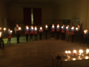 Blurry image of Guides standing in a horseshoe formation all holding candles. At the opening of the horsehow there is a table with coloured candles lit.
