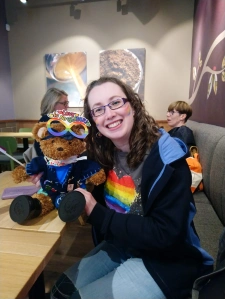 Woman with brown curly hair and glasses wearing a charcoal grey t-shirt with a rainbow pride heart design, with a girlguiding hoodie over the top. She is holding a brown bear wearing wellies and a poncho blanket covered in badges and some pride glasses.